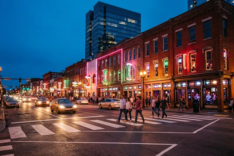Lower Broadway in Nashville at blue hour, with brick honky-tonks lit in neon and people crossing.